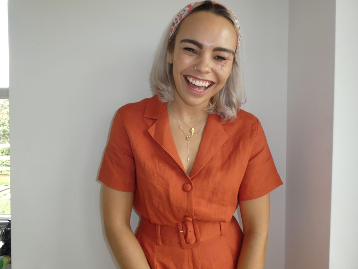 A woman with blonde hair stands in front of a blank wall. She is wearing orange and smiling at the camera.