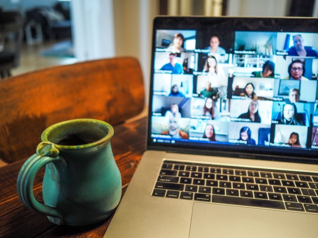 A laptop showing a Zoom video call, with lots of faces on screen, and a mug next to the laptop on the desk.